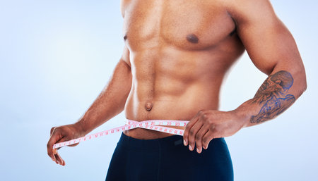 Black man, fitness and body, measuring tape and abs with health, weight loss and active lifestyle on blue background. Shirtless male bodybuilder, exercise and wellness with diet, healthy and strongの写真素材
