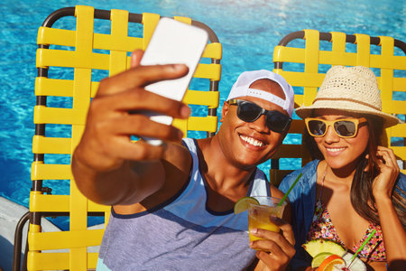 One for the photo album. High angle shot of an affectionate young couple taking selfies while enjoying a few drinks poolside.の写真素材