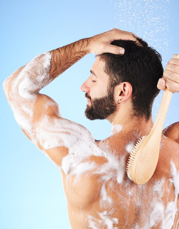 Shower, cleaning and back of man with water splash, brush and soap for wellness, hygiene and grooming. Skincare, self care and happy male with foam, bath cosmetics and washing body on blue backgroundの写真素材