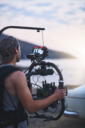 The light is fading. Rearview shot of a focused young man shooting a scene with a state of the art video camera outside on a beach during the day.の写真素材