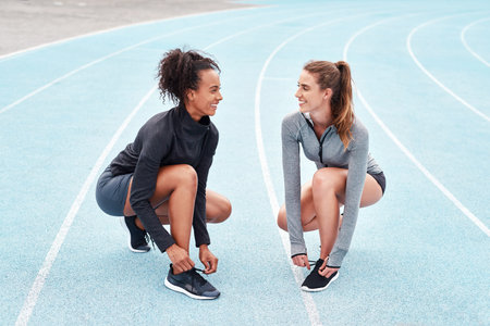 Teamwork is the best motivation. Full length shot of two attractive young athletes crouching together and tying their shoelaces before running on a track.の写真素材