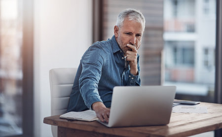 Working through ways to better manage his business. a thoughtful mature businessman working on a laptop in an office.の写真素材