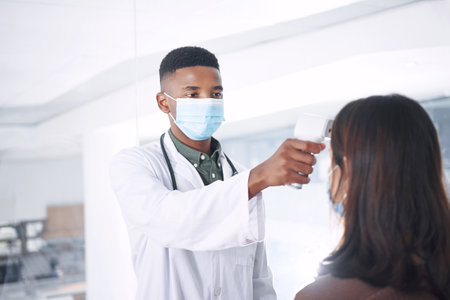 This will only take a second. a young doctor wearing a mask and standing with a patient in the clinic while taking her temperature.の写真素材