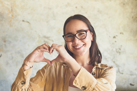 Choose kindness and spread happiness. Portrait of a young woman making a heart shape with her hands against a wall.の写真素材