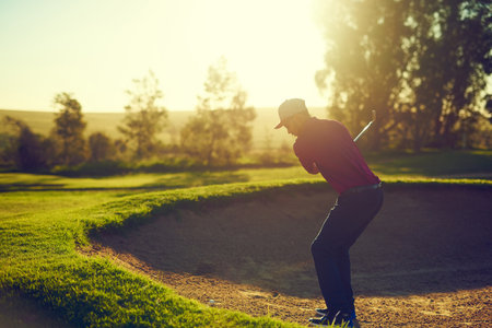 Hes the boss of that bunker. a young man hitting the ball out of the bunker during a round of golf.の写真素材