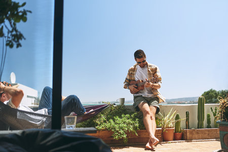Perfect song for a perfect day. a cheerful young man playing on his guitar while his friend listens outside on a balcony during the day.の写真素材