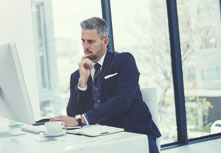 The hard work responsible for his career success. a mature businessman using a computer at his desk in a modern office.の写真素材