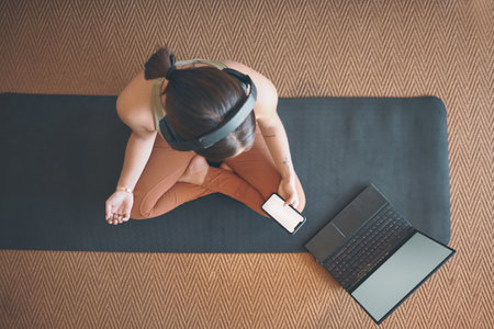 Learning from the yoga masters online. High angle shot of a young woman using a cellphone and laptop while exercising at home.の写真素材