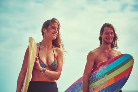Surfing brings us closer together. a young couple walking on the beach with their surfboards.の写真素材