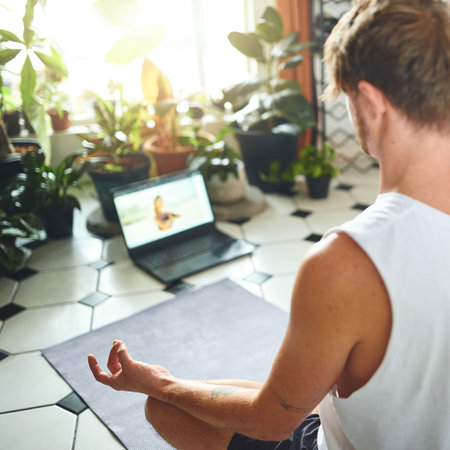 Yoga class brought straight to you. a young man using a laptop while meditating in the lotus position at home.の写真素材