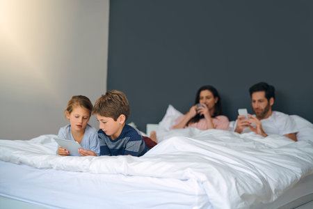 Its too early to do anything therefor were chilling. an adorable little girl and boy using a tablet together while their parents are chilling in bed at home.の写真素材