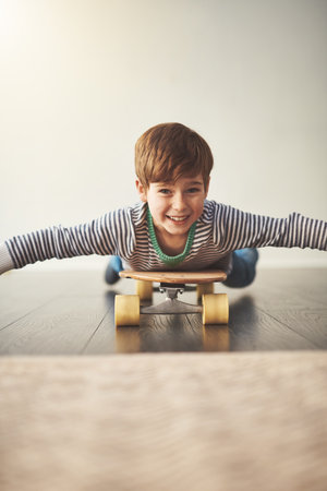 I dont need to stand on a skateboard to have fun. Portrait of a cheerful little boy lying down on a skateboard at home during the day.の写真素材