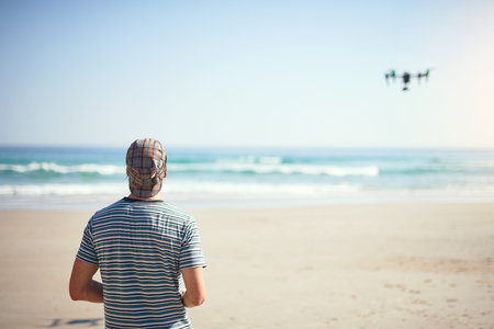 We have liftoff. a focused young man holding a remote control to fly a drone on a beach outside...