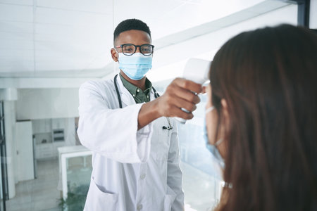 Im strict about Covid rules and regulations. a young doctor wearing a mask and standing with a patient in the clinic while taking her temperature.の写真素材