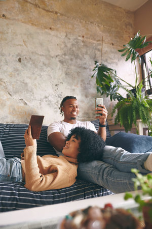 Together in their own worlds. a young woman reading a book while her boyfriend uses a cellphone at home.の写真素材