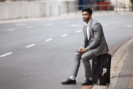 Suitcase, road and businessman in the city for travel to airport for corporate company trip. Phone, bag and professional male employee sitting in urban town road waiting for a cab, taxi or transport.の写真素材