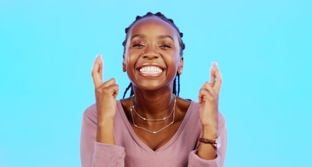 Face, fingers crossed and black woman with smile, hope and motivation with lady against blue studio...