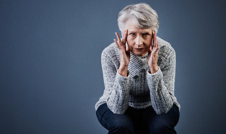 She can read your thoughts. Studio shot of a elderly woman sitting with her hands touching the sides of her head while looking at the camera.の写真素材