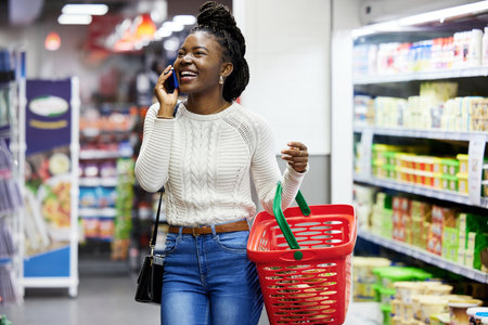 Happy black woman, supermarket and shopping with phone call, choice or discount in funny chat, customer experience. Plastic basket, grocery store and mall for retail, talking and girl with smartphoneの写真素材