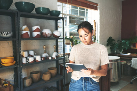 I should be able to slot you in todays schedule. Cropped portrait of an attractive young business owner standing alone in her pottery studio and using a tablet.の写真素材