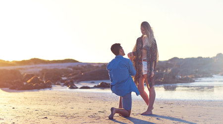 Theres something Ive been wanting to ask you. a young man holding his girlfriends hand on bended knee at the beach.の写真素材
