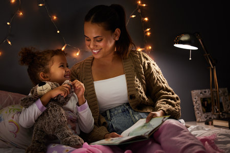 Night, book and learning with mother with daughter in bedroom for storytelling, fantasy and smile. Education, fairytale and love with woman reading to girl in family home for bedtime, happy or relaxの写真素材