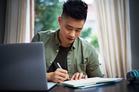 Working out a rough plan. a handsome young man taking notes while working on his laptop at home.の写真素材