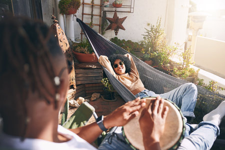 Enjoying the relaxing beats. a young woman relaxing on a hammock while listening to her boyfriend play drums.の写真素材