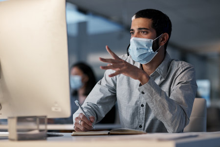 Call center, man and communication with mask at computer while writing notes for advice, customer service and sales. Male telemarketing agent, virus protection and desktop for consulting questionsの写真素材
