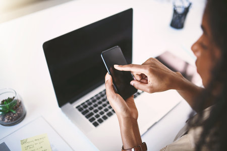 Making success happen with smart apps. a young businesswoman using a mobile phone and laptop at her desk in a modern office.の写真素材