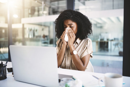 This runny nose is going to ruin my day. a young businesswoman blowing her nose with a tissue while working in a modern office.の写真素材