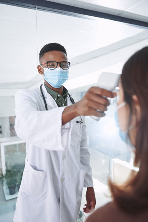 Lets check that temperature. a young doctor wearing a mask and standing with a patient in the clinic while taking her temperature.の写真素材