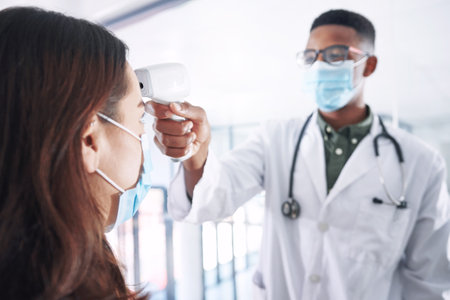 A temperature check is a must. a young doctor wearing a mask and standing with a patient in the clinic while taking her temperature.の写真素材