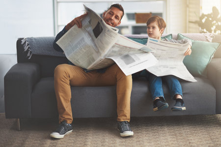 What does the paper say. a carefree little boy and his father reading the newspaper while being seated on the sofa at home during the day.の写真素材