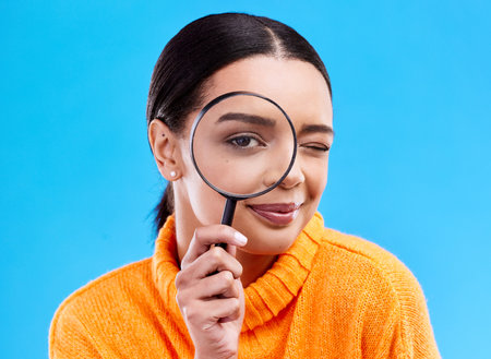 Search, portrait and female with a magnifying glass in a studio for an investigation or detective cosplay. Happy, smile and headshot of a woman model with a zoom magnifier isolated by blue backgroundの写真素材