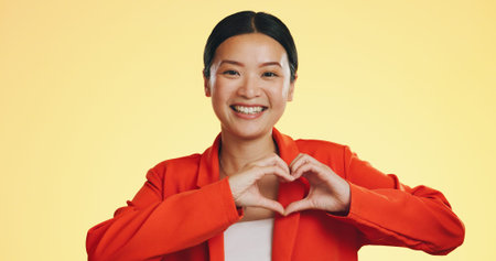 Smile, heart in hands and face of Asian woman on yellow background for love, support and charity...