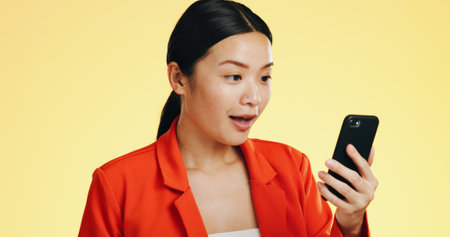 Winner, success celebration and Asian woman with phone in studio isolated on a yellow background. Surprise, fist pump or happy female with mobile to celebrate after winning lottery prize or good newsの写真素材