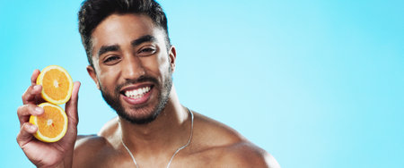 Beauty, orange and face of man in studio for skincare, wellness and citrus treatment on blue background. Fruit, facial and portrait of indian male model excited for organic vitamin c skin cosmeticsの写真素材