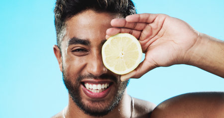 Skincare, lemon and face of man in studio for beauty, wellness and citrus treatment on blue background. Fruit, facial and portrait of indian guy model relax with organic, anti aging or skin cosmeticsの写真素材