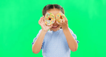 Donut on eyes, smile and child on green background with cake over face for funny, meme and comic. Food, excited kid and isolated happy girl with sweets, dessert treats and sugar doughnuts in studioの写真素材