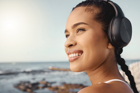 Headphones, fitness and happy woman at beach for exercise, vision or running mindset, goals or music ideas. Health, podcast and athlete, runner or sports person thinking and listening to audio by seaの写真素材