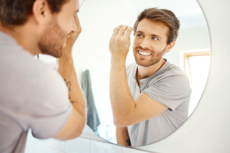 Man, tweezer and eyebrow in bathroom mirror with smile, self care and hygiene to start morning in home bathroom. Young guy, person and tool for beauty, facial cosmetics and wellness for hair removalの写真素材