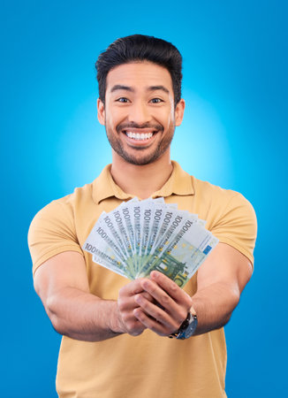 Money, portrait and man or winner with bonus offer, financial success and winning, finance loan or lottery fan. Young asian person with savings, cash and profit isolated on studio, blue backgroundの写真素材