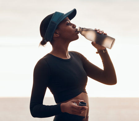 Drinking water, fitness and black woman by ocean for exercise, marathon training and running. Sports, hydration and female person with liquid in bottle for thirst, cardio workout and body wellnessの写真素材