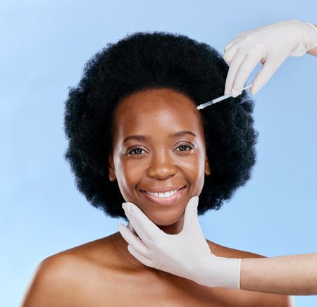 Black woman, face injection and beauty with cosmetic procedure, fillers and dermatology on blue background. Liquid collagen, facial and skincare with hands, female model and smile in portraitの写真素材