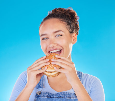 Burger, smile and woman eating fast food and happy with lunch meal with a smile isolated in a studio blue background. Breakfast, craving and portrait of young female person enjoy snack or sandwichの写真素材