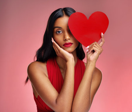 Woman, paper heart and face in studio portrait for makeup, beauty or romantic sign by red background. Girl, fashion model and cardboard emoji with love for cosmetic, valentines day date and aestheticの写真素材