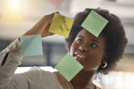 Black woman, writing or entrepreneur brainstorming ideas on glass board with startup strategy in office. Face, sticky notes or businesswoman planning a schedule, timeline or tasks for company growthの写真素材