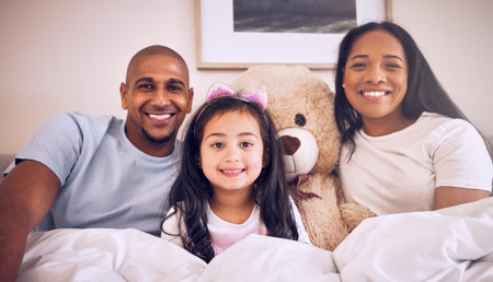 Family, morning and happy portrait on a bed at home with a smile, teddy bear and comfort for quality time. Man, woman or parents and a girl kid together in the bedroom for bonding with love and careの写真素材