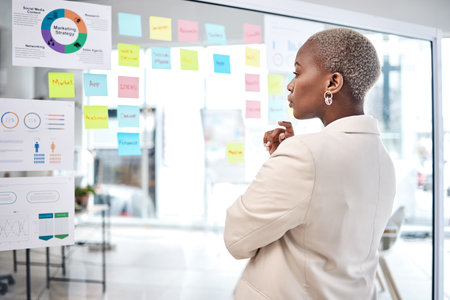 Black woman, thinking and glass board for project management, marketing or planning at office. African female person, manager or employee checking project plan, tasks or statistics at the workplaceの写真素材